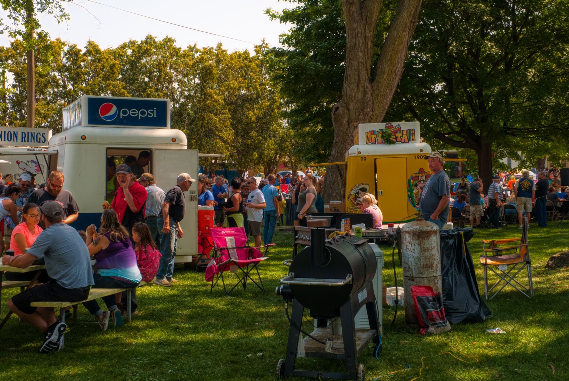 Crowded outdoor food festival under lush trees with people enjoying food stalls.