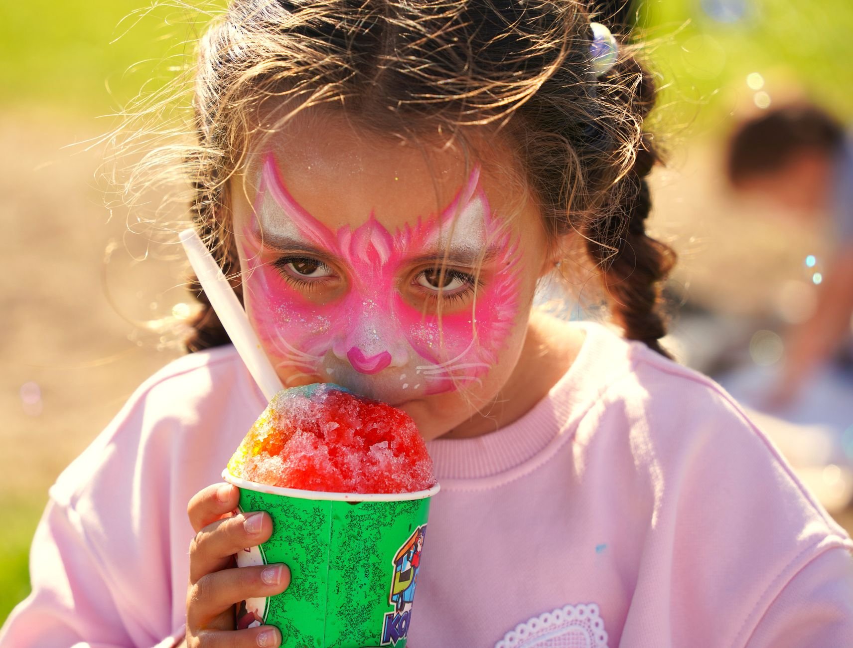 Child with pink face paint and braided hair enjoys a colorful sno-cone outdoors in bright sunlight.