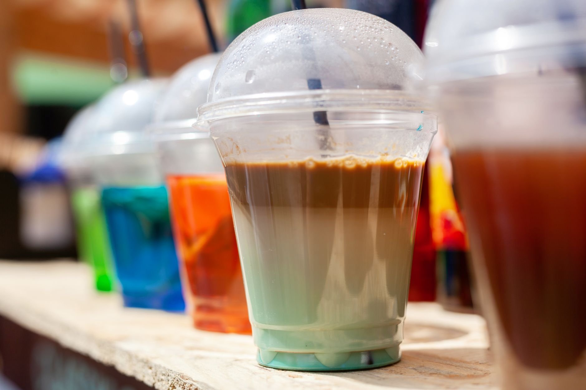Close-up of vibrant colorful iced drinks in plastic cups with straws, on a sunny day outdoors.